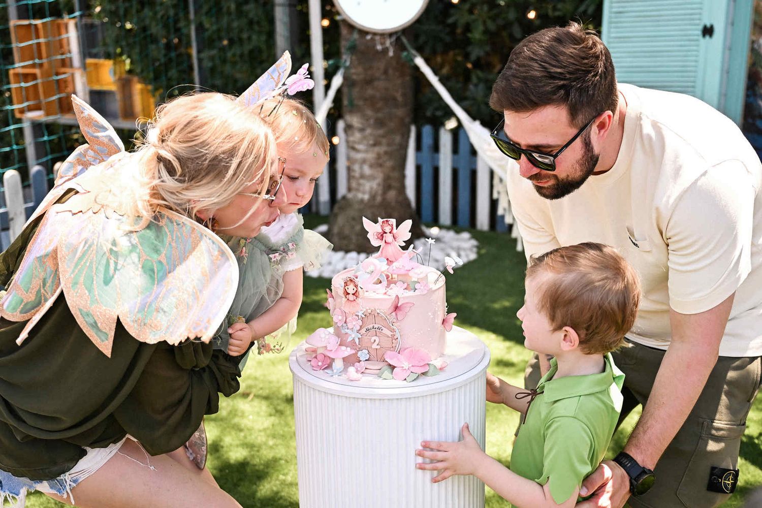 Parents and their children leaning in together to blow out the candle on a beautifully decorated birthday cake outdoors.