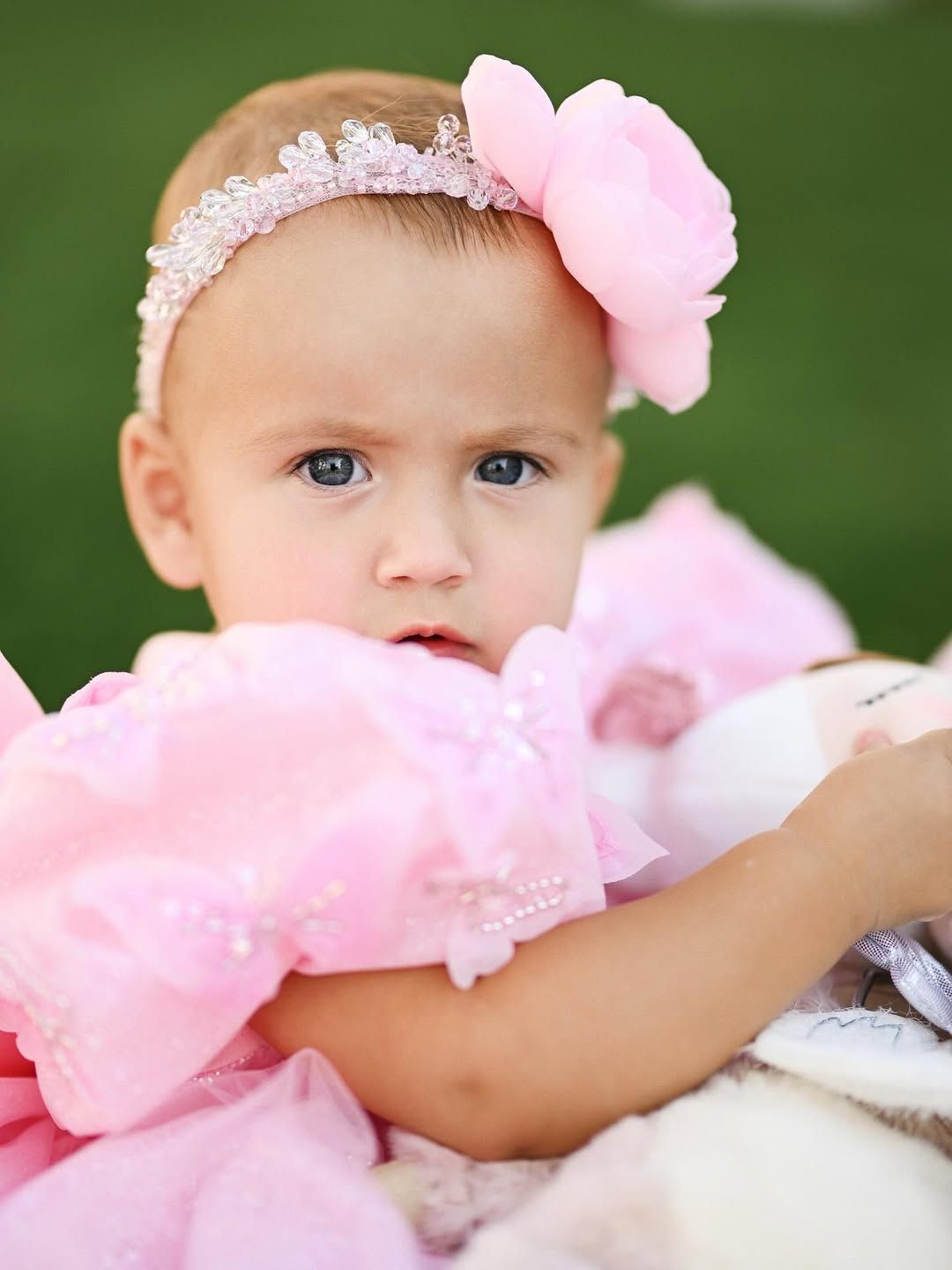Beautiful close-up portrait of a baby girl with blue eyes wearing a pink dress and a floral crystal headband.