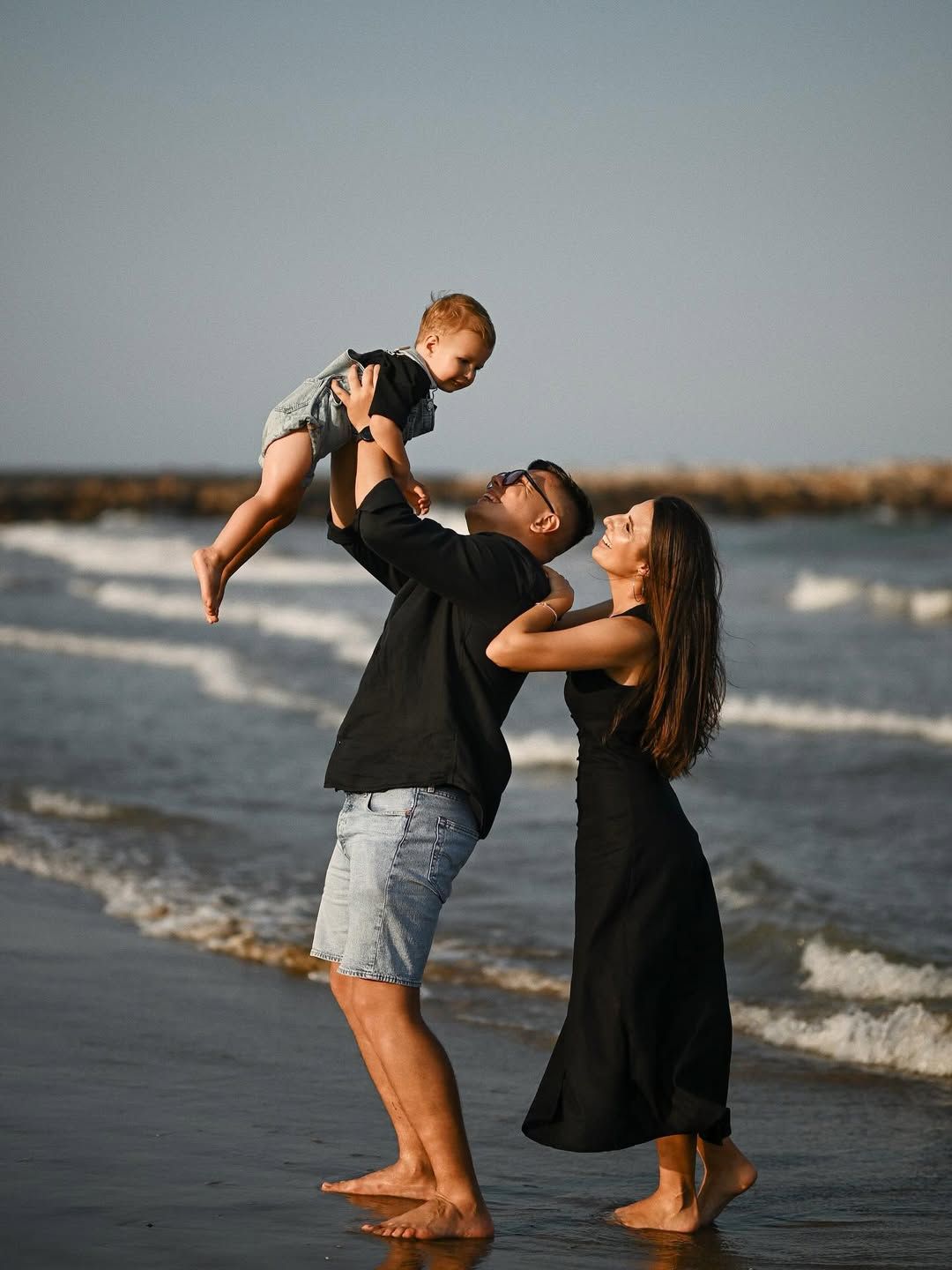 A father lifts his little son above his head while the mother hugs him on the sandy beach of Port Saplaya at sunset