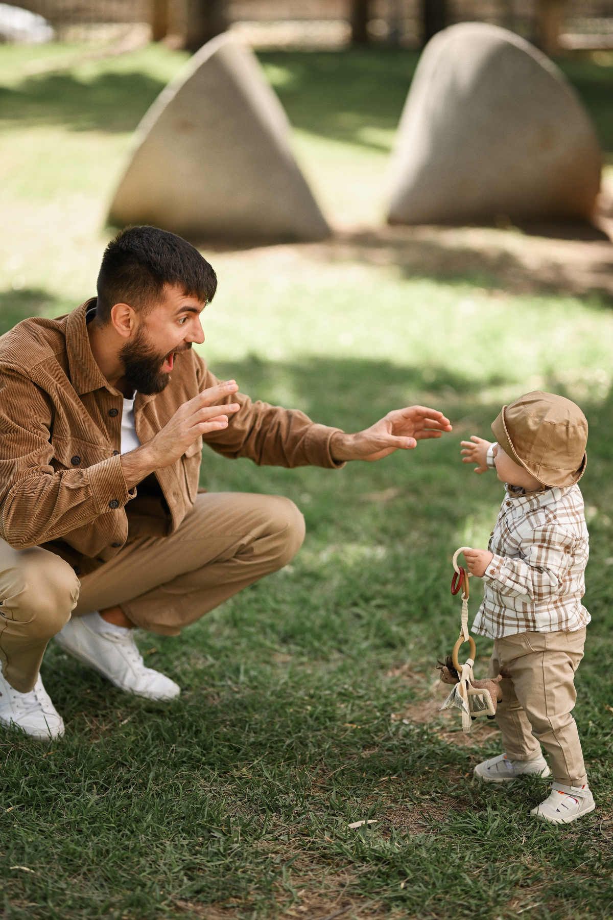 Dad crouching on the green grass with open arms, cheering for his toddler son taking steps in Viveros Park, Valencia.