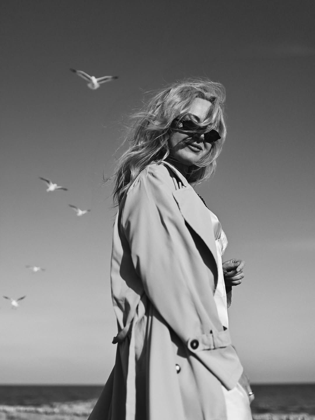 Artistic black and white portrait of a blonde woman in sunglasses looking over her shoulder with seagulls flying in Costa Blanca.