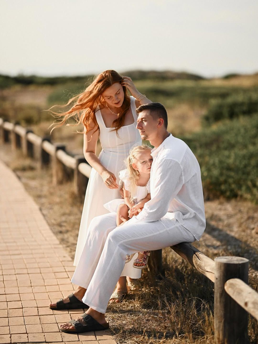 Mother with her daughter and father sitting on a rustic wooden fence in a nature park during a sunset shoot.
