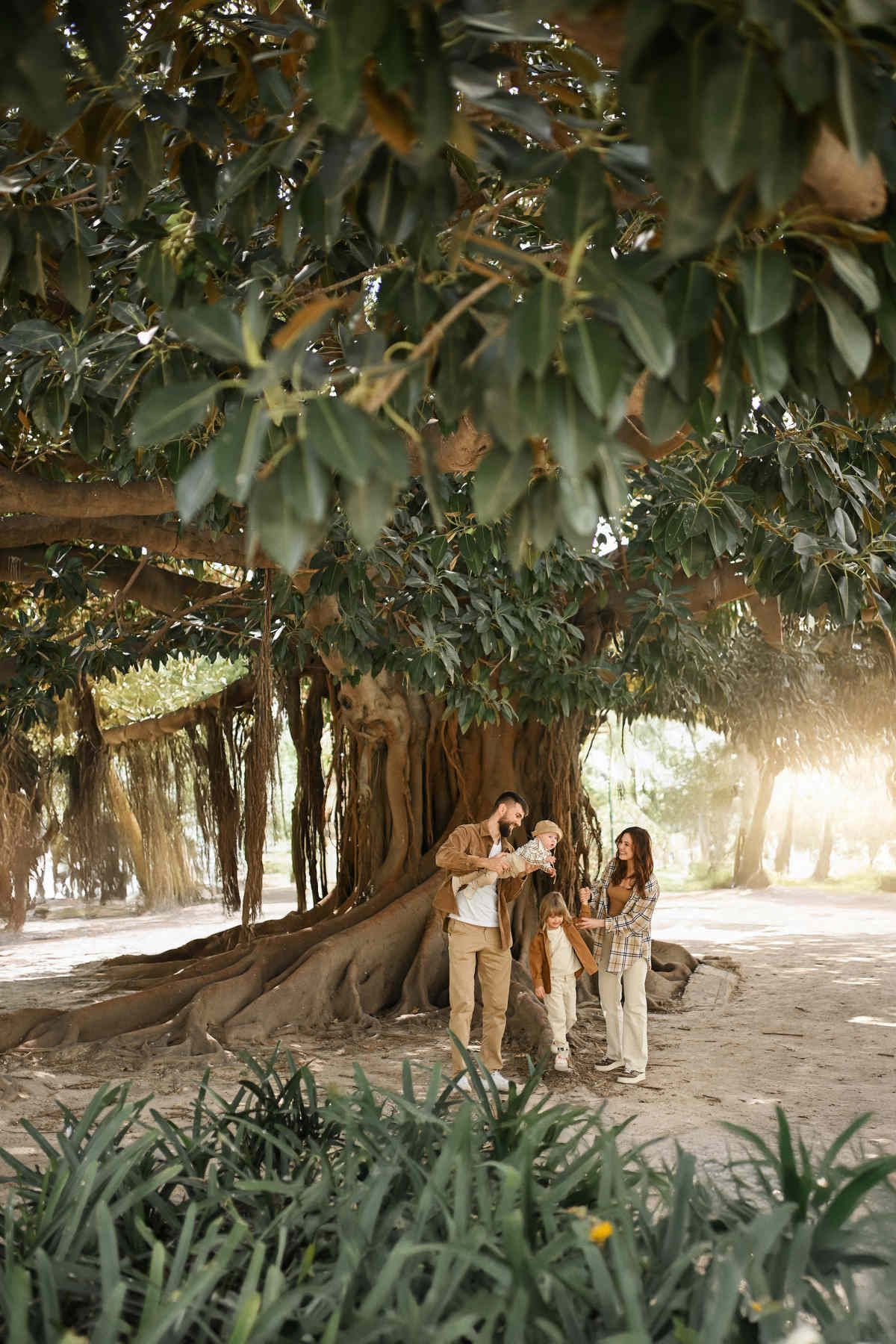A happy family with two young children enjoying an outdoor photo session beneath the expansive branches of a giant, ancient tree in Valencia