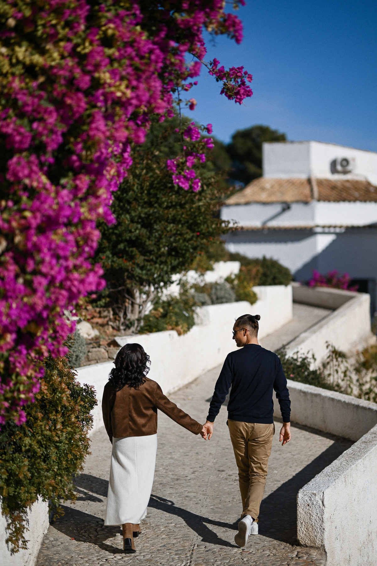 A pregnant couple holding hands and walking up a picturesque path surrounded by white architecture and flowers in Altea