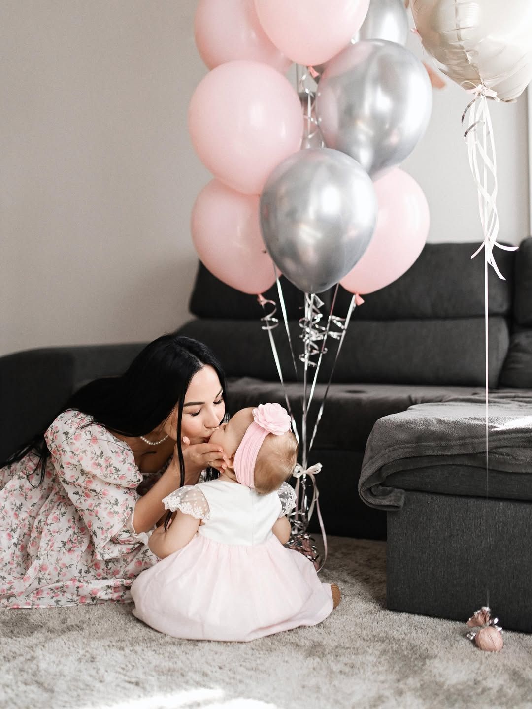Loving mother kissing her baby daughter next to pink and silver helium balloons on the living room floor.