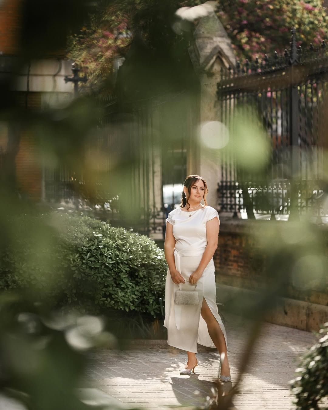 Portrait of a stylish woman framed by green foliage, standing in a quiet historic plaza during a Valencia photoshoot