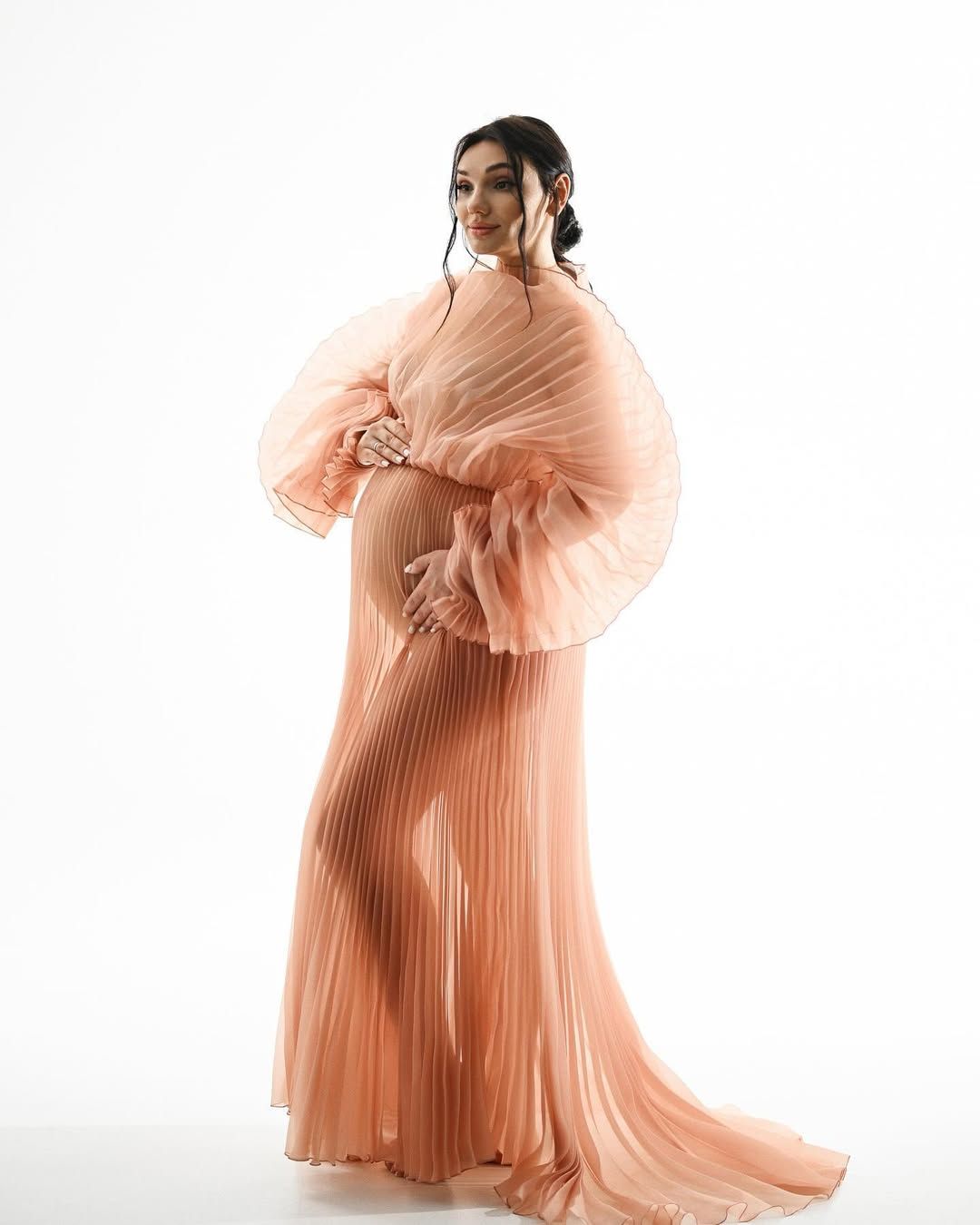 Pregnant woman in a flowing peach pleated dress posing during an elegant studio maternity photoshoot in Valencia