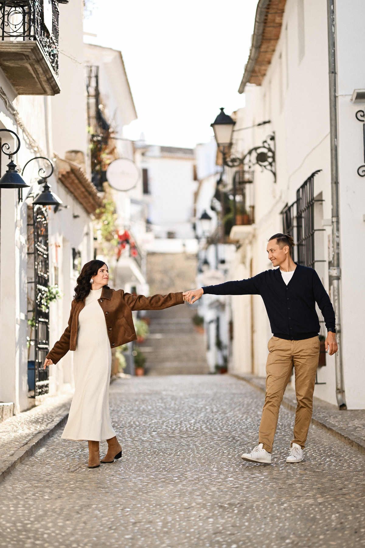 A happy expectant couple holding hands and walking playfully down a historic cobbled street in Altea Old Town