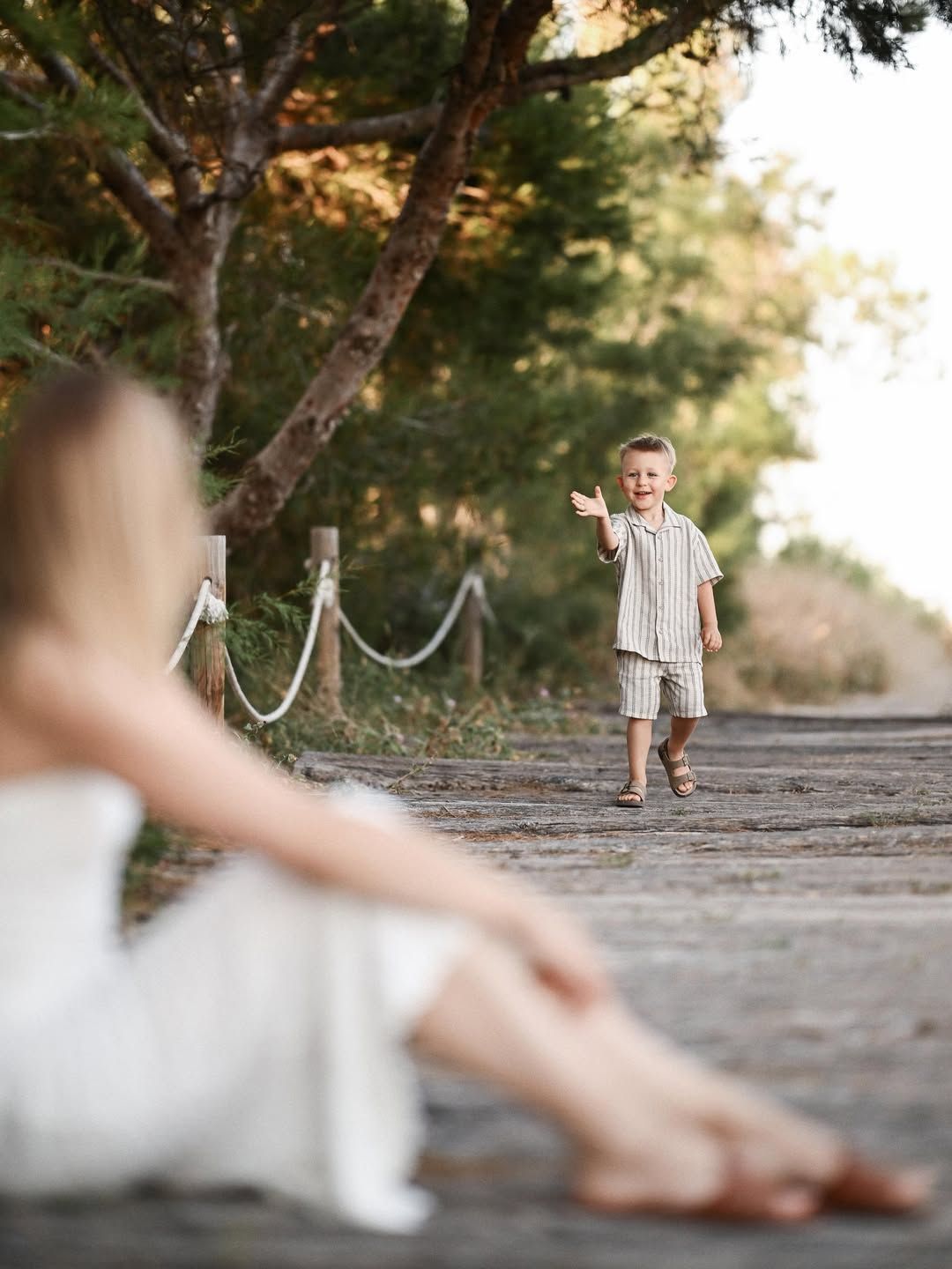 A little boy in a striped outfit walks along a wooden path reaching out to his mother at El Saler pine forest.