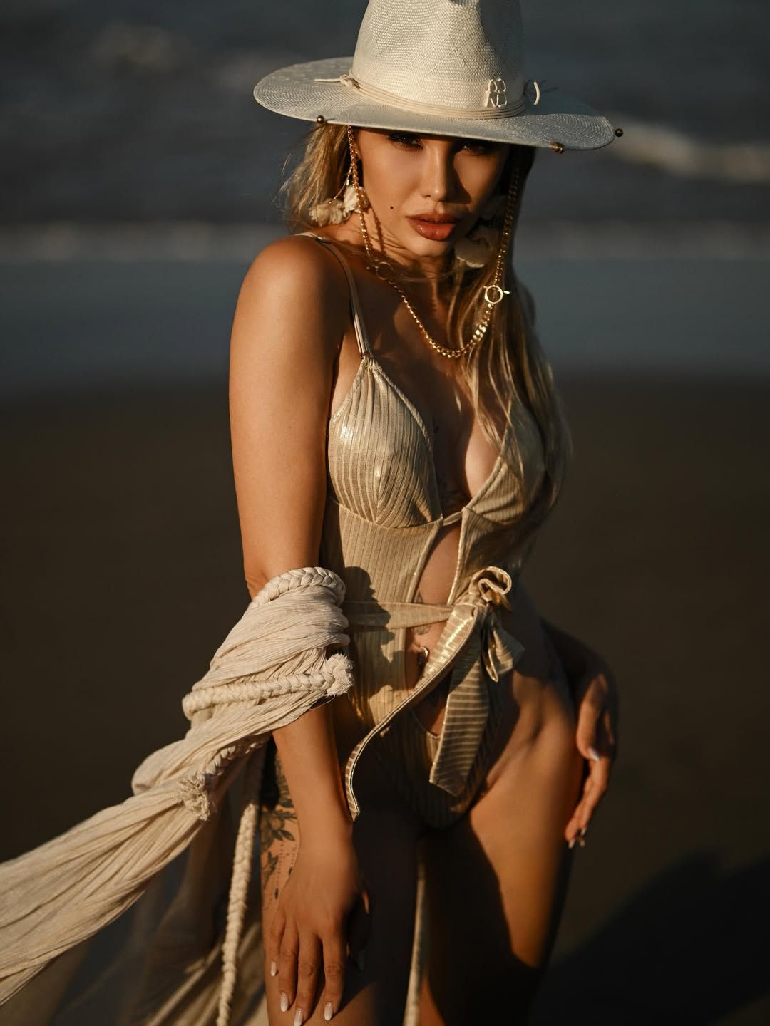 An editorial portrait of a woman in a gold swimsuit and white hat standing by the sea shore, perfect for a social media update.