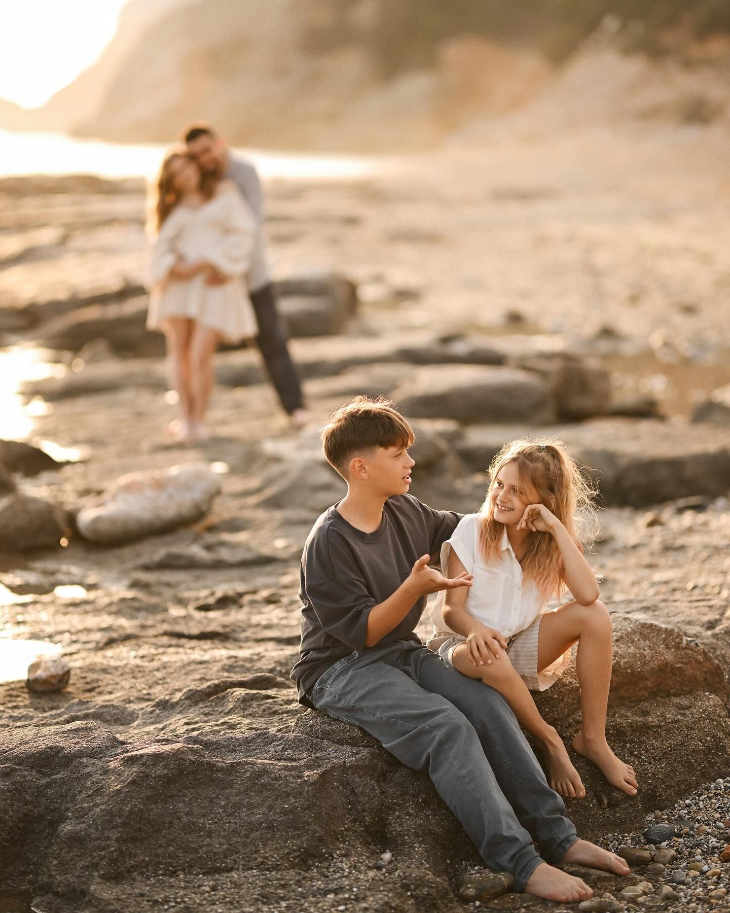 A young boy talking to his sister while sitting on a rocky beach, with parents blurred in the background in Costa Blanca.