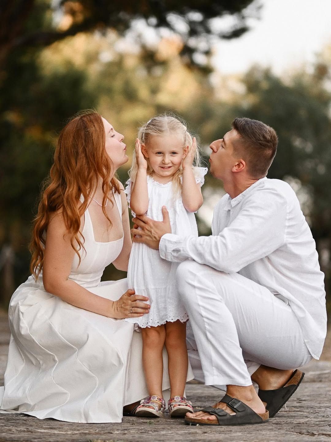 Parents playfully kissing their daughter's cheeks while she holds her ears in an outdoor El Saler park location.