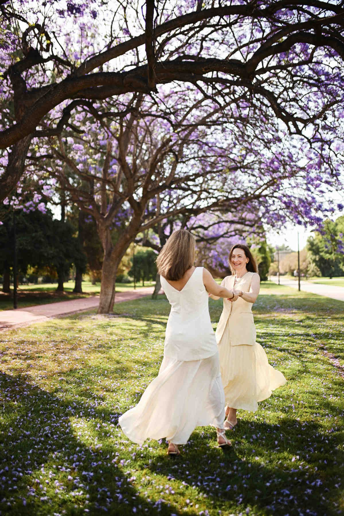 Two sisters holding hands and spinning joyfully on a green lawn covered in fallen purple blossoms.