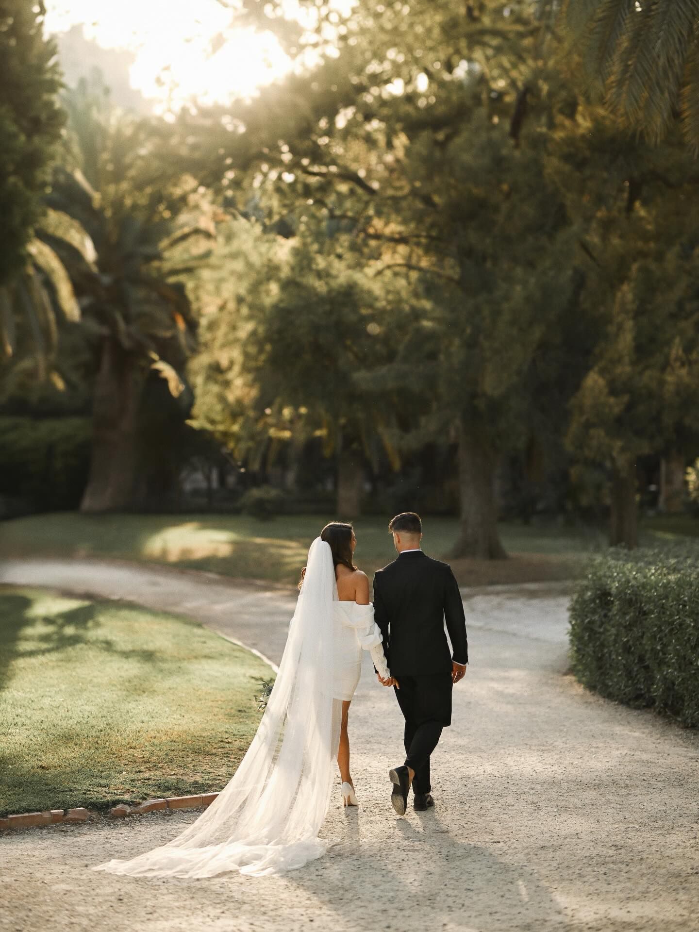A bride in a short dress with a long veil and a groom in a black suit walk down a path in a sun-drenched Valencia park.