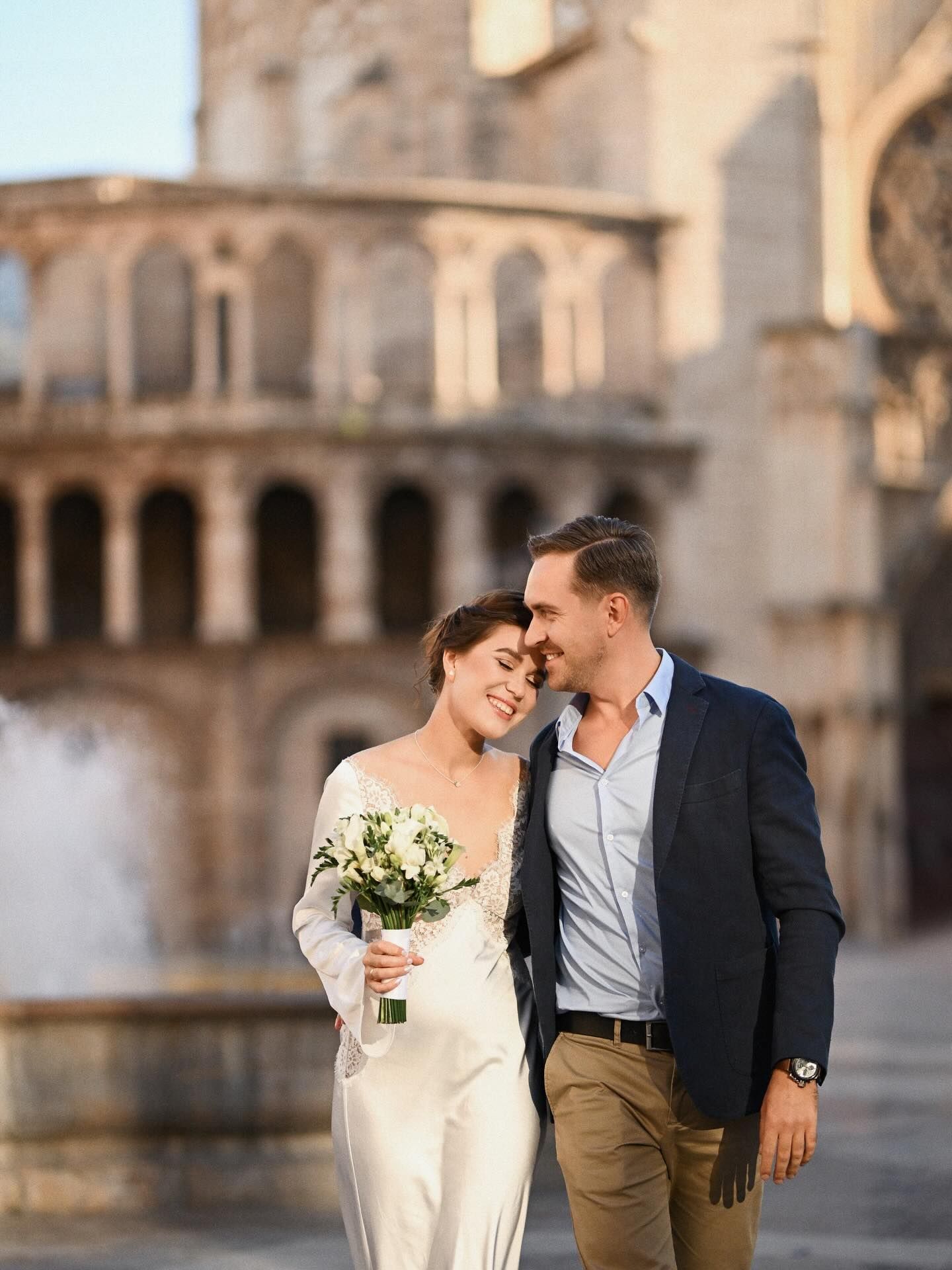 A laughing newlywed couple walking past a historic fountain and cathedral in the center of old town Valencia during golden hour.