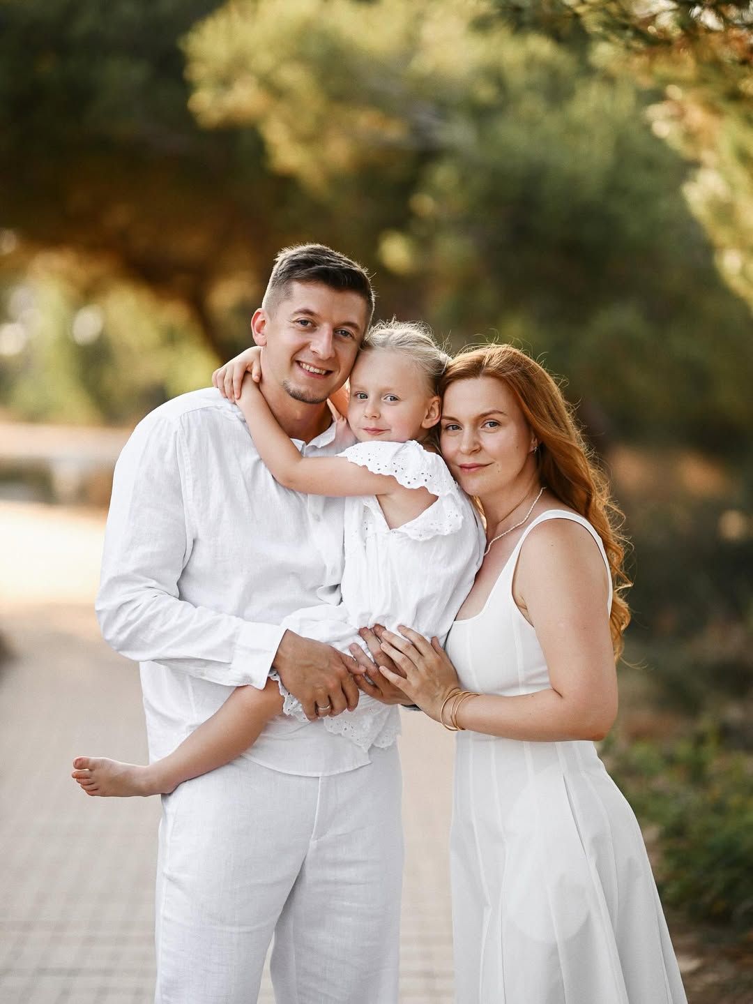 Mom, dad, and daughter in matching white outfits hugging against a pine forest backdrop in El Saler Nature Park.