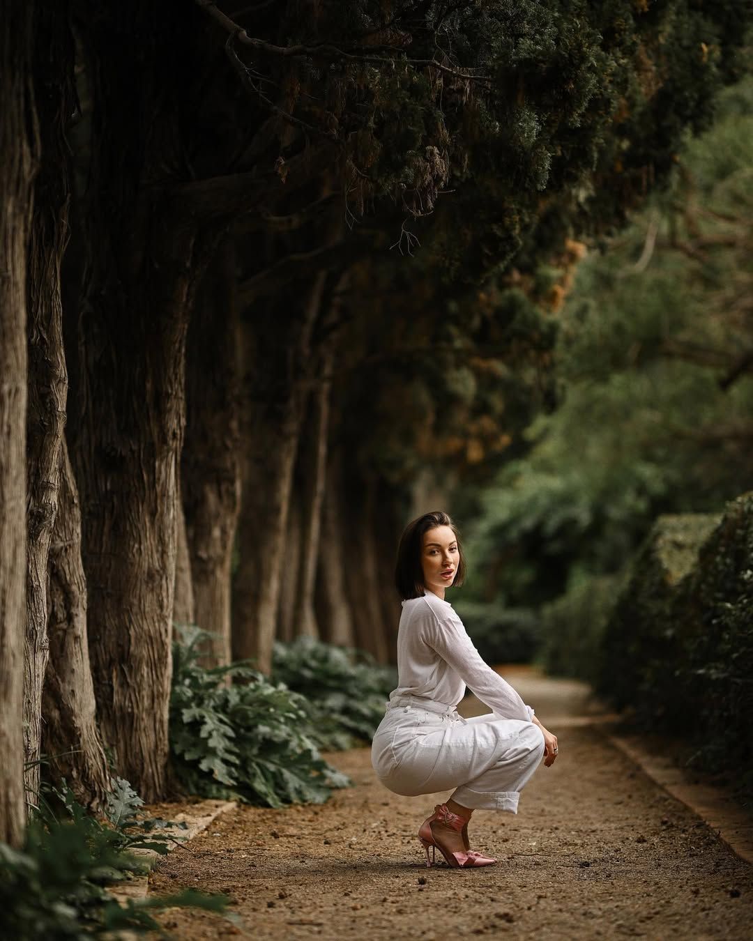 A woman in a white casual outfit posing on a dirt pathway under massive green trees in Monforte Gardens.