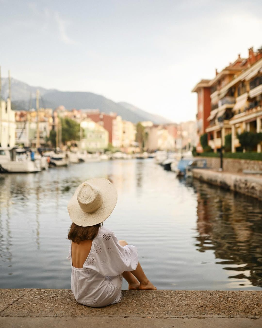 A woman in a sun hat sits with her back to the camera, overlooking the colorful buildings, boats, and canals of Port Saplaya