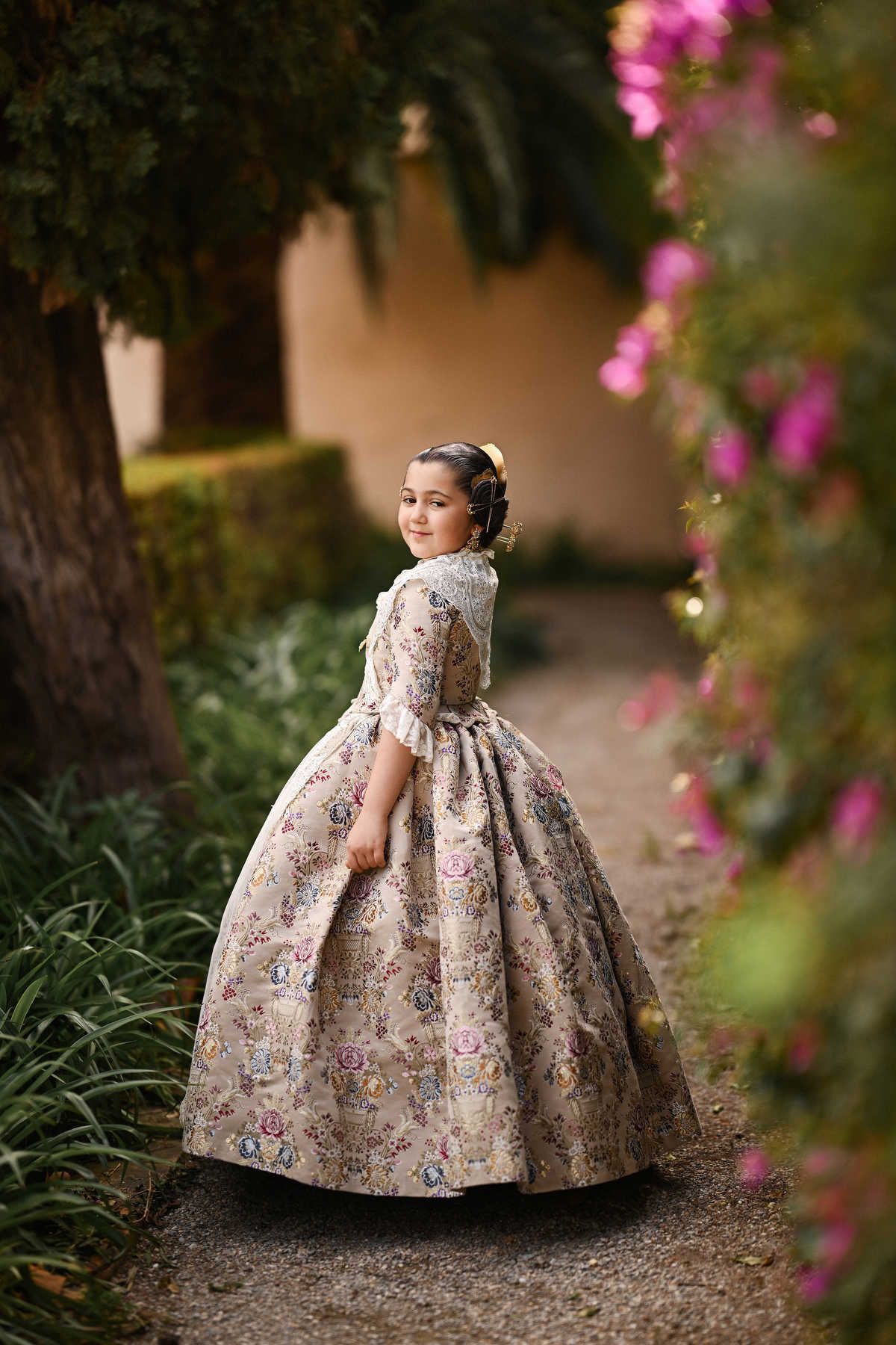 A graceful full-body portrait of a young girl posing in her intricate traje de fallera against the lush greenery of Jardines de Monforte.