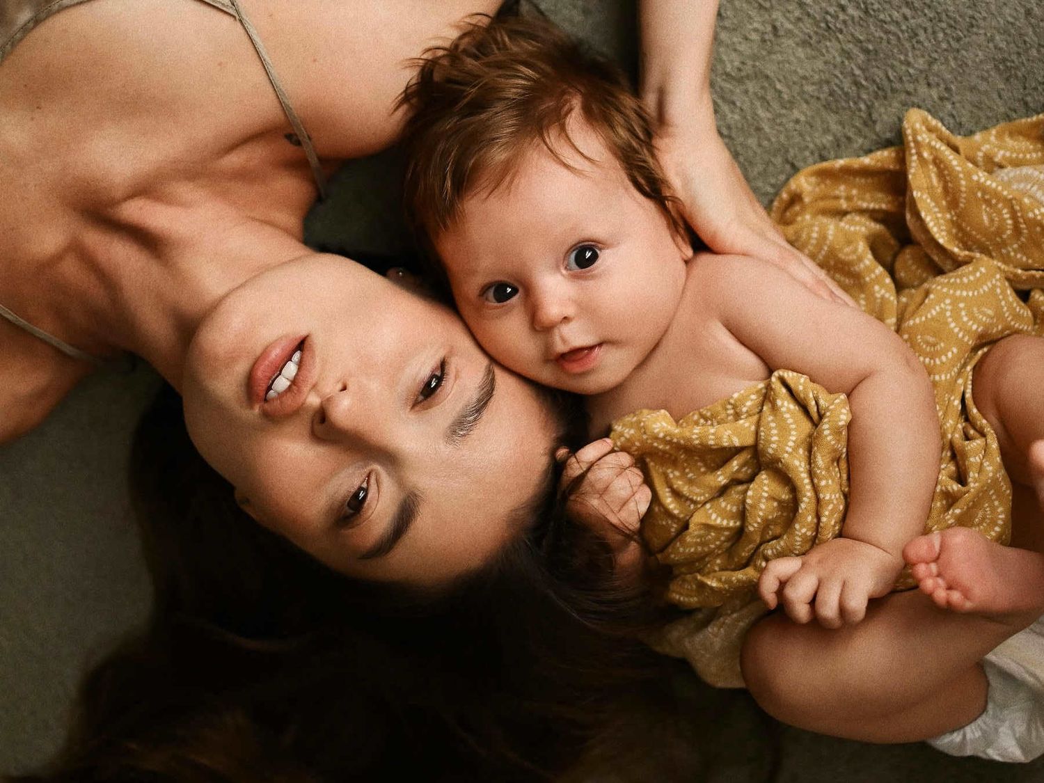A peaceful portrait of a mother and her baby lying together on the floor, looking into the camera with an emotional bond.