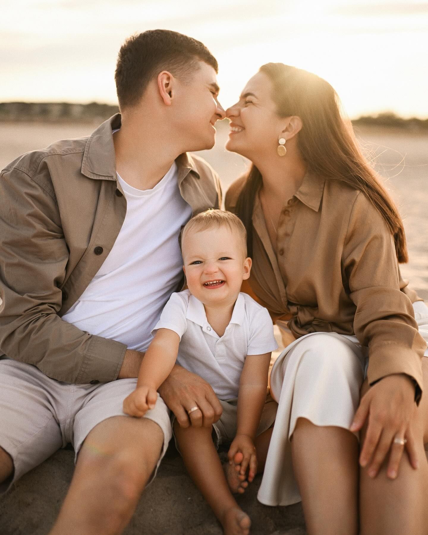 Close-up portrait of a joyful family sitting on the beach during golden hour, with their little child smiling at the camera.