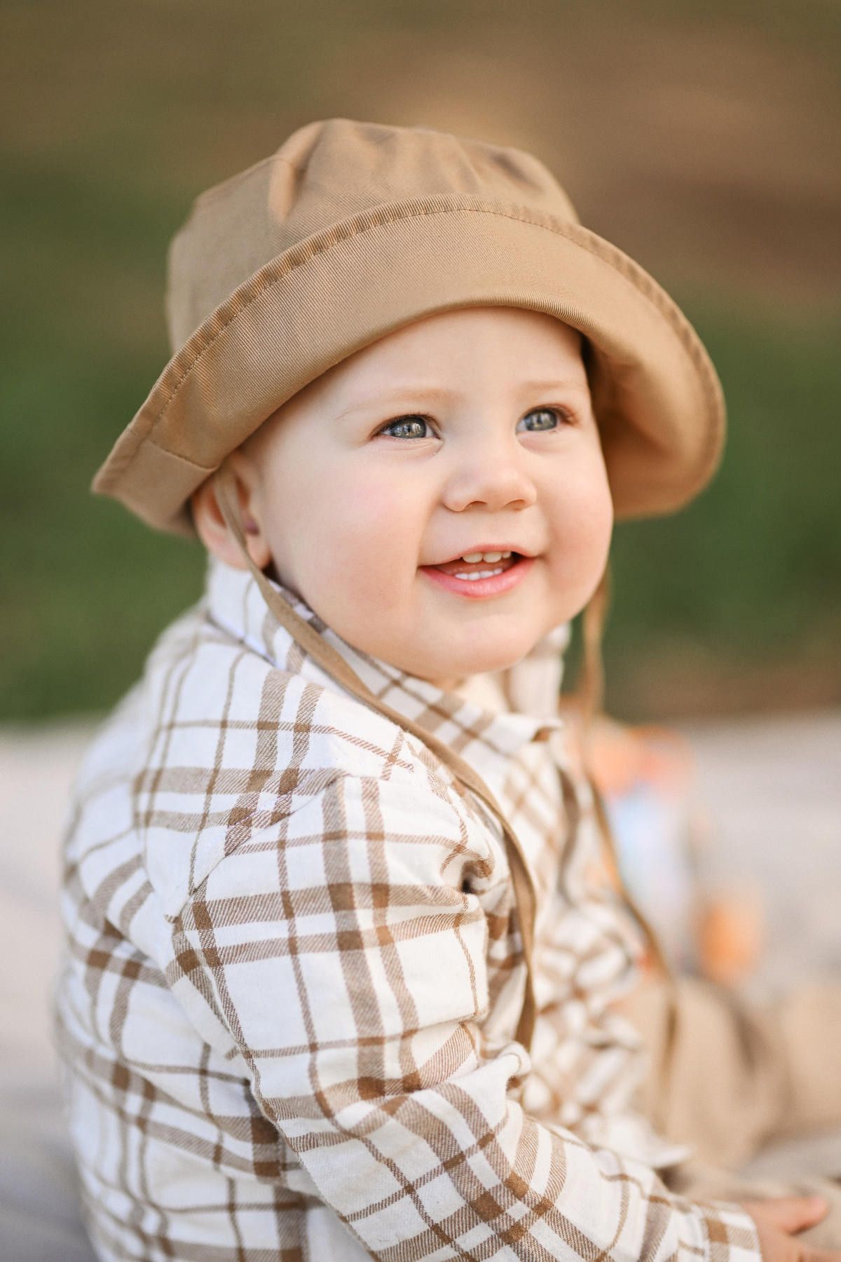 Close-up portrait of a smiling toddler boy in a beige hat sitting on the grass during a family session in Viveros Park.