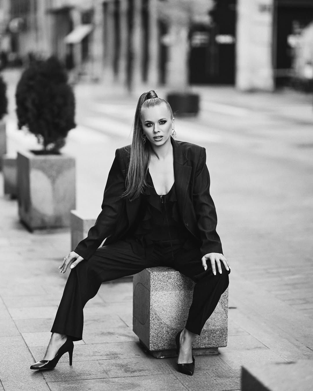 A high-fashion black and white portrait of a model in a tailored suit, sitting confidently on a stone urban block in Valencia.