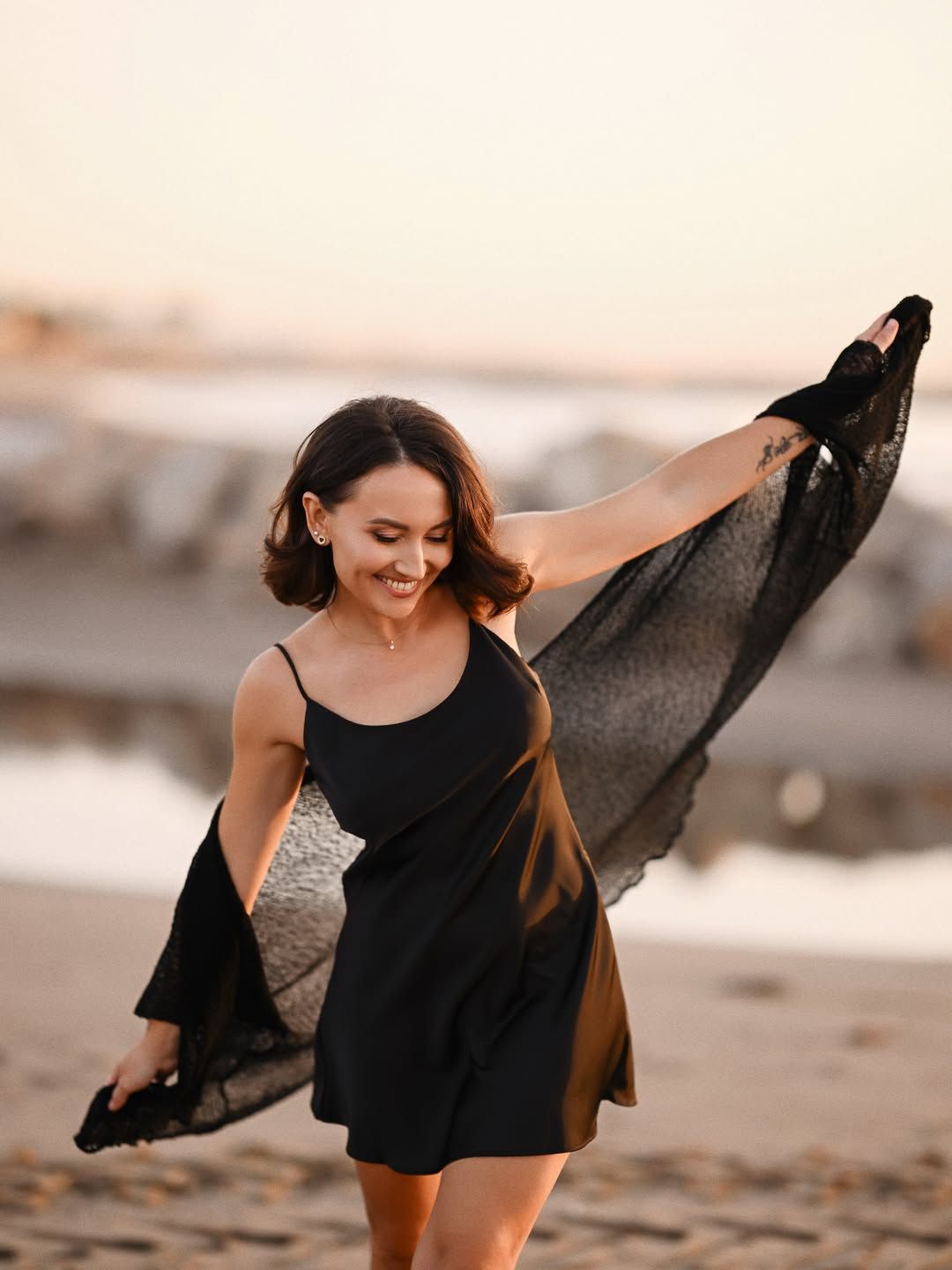 Lifestyle women's portrait in Valencia, smiling woman in a black dress throwing a sheer dark fabric into the sea breeze.