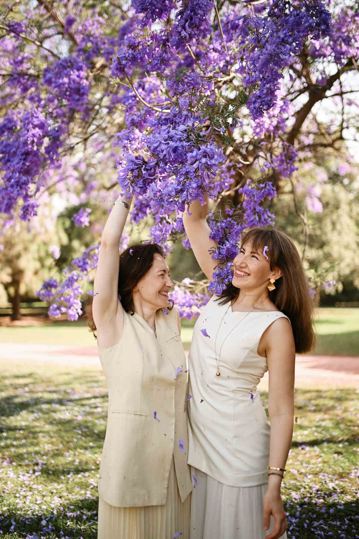 Two joyful women playing under Jacaranda branches, with falling purple petals caught mid-air around them.