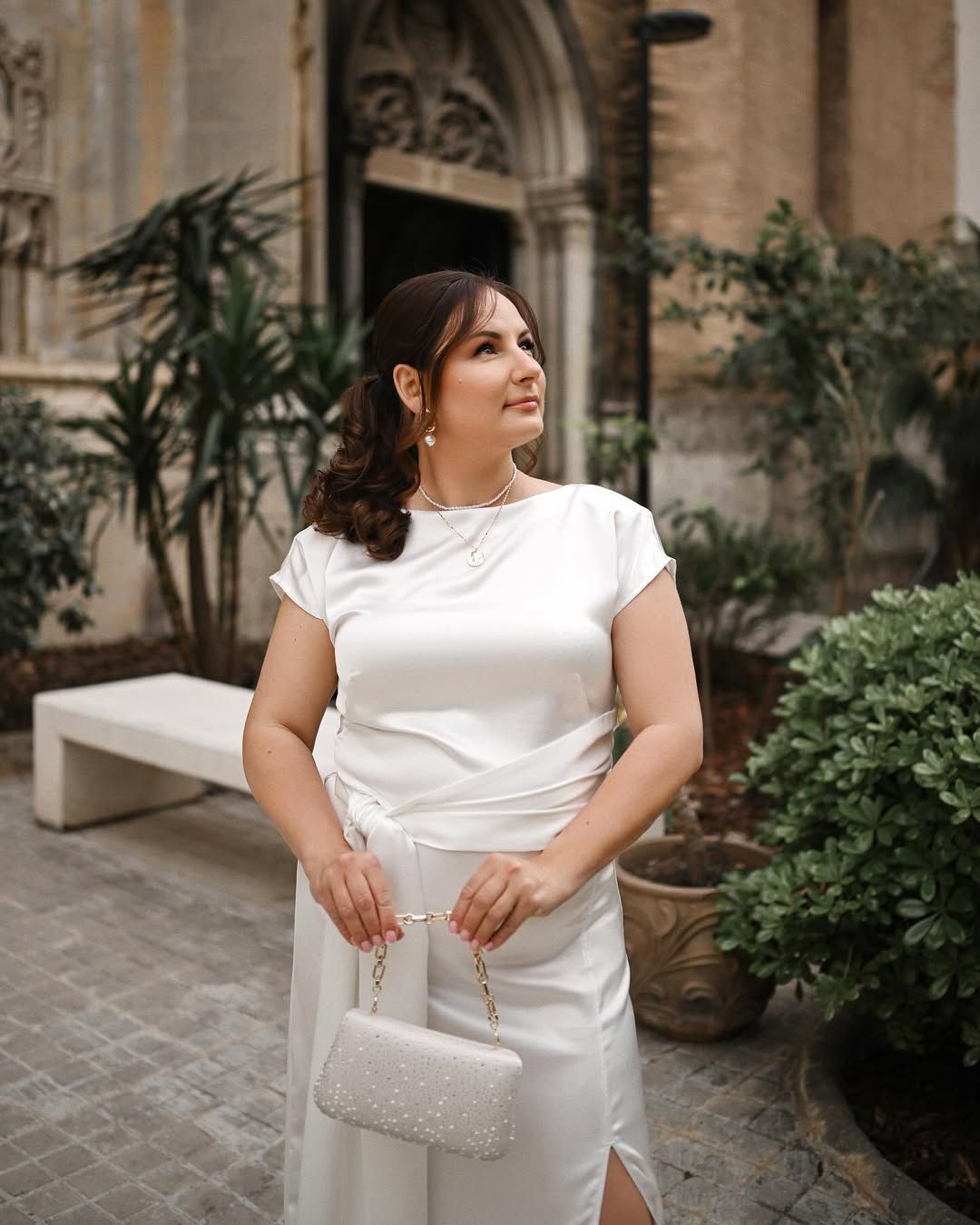Beautiful woman in a white dress holding a clutch bag in a tranquil urban garden setting in Valencia