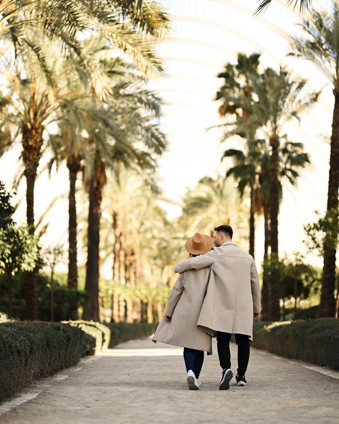 Couple walking arm-in-arm down a path lined with tall palm trees at the Umbracle garden during a love story session.