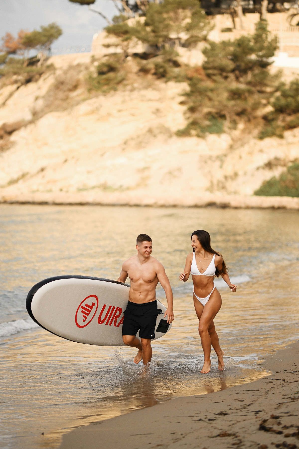 A happy couple running along the beach with a paddleboard during a relaxed lifestyle photoshoot in Moraira