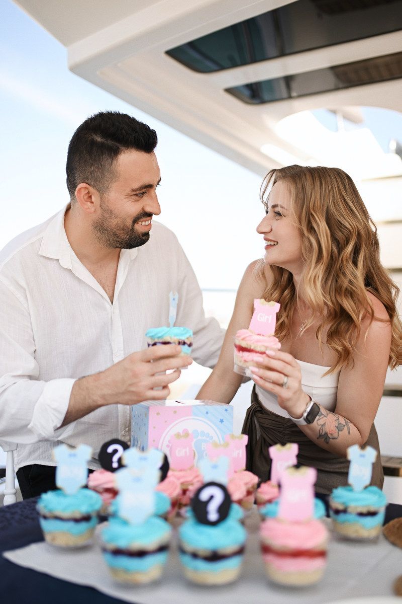 Happy parents-to-be smiling at each other next to a styled gender reveal candy bar on a Mediterranean yacht.