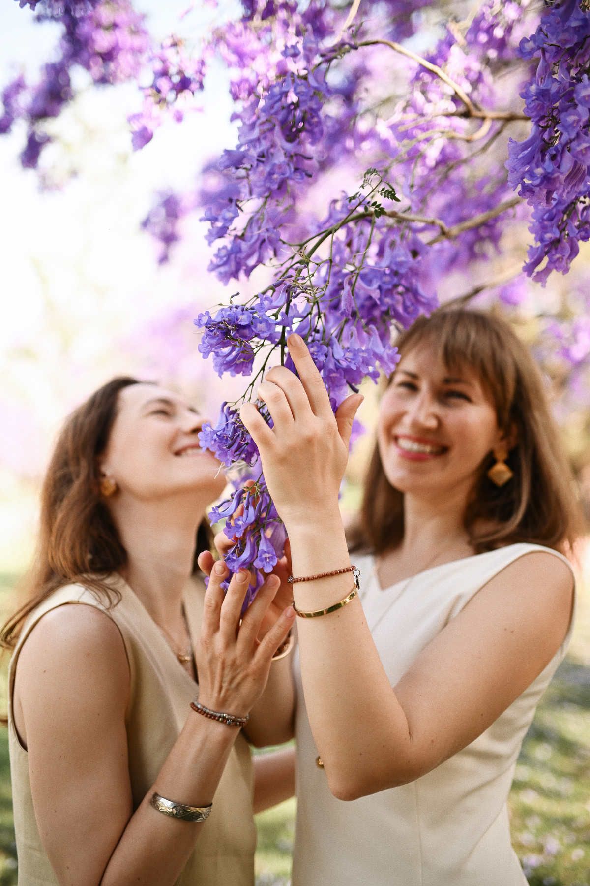Stylized duo portrait of two women gently touching and admiring vibrant purple Jacaranda blossoms in a park.