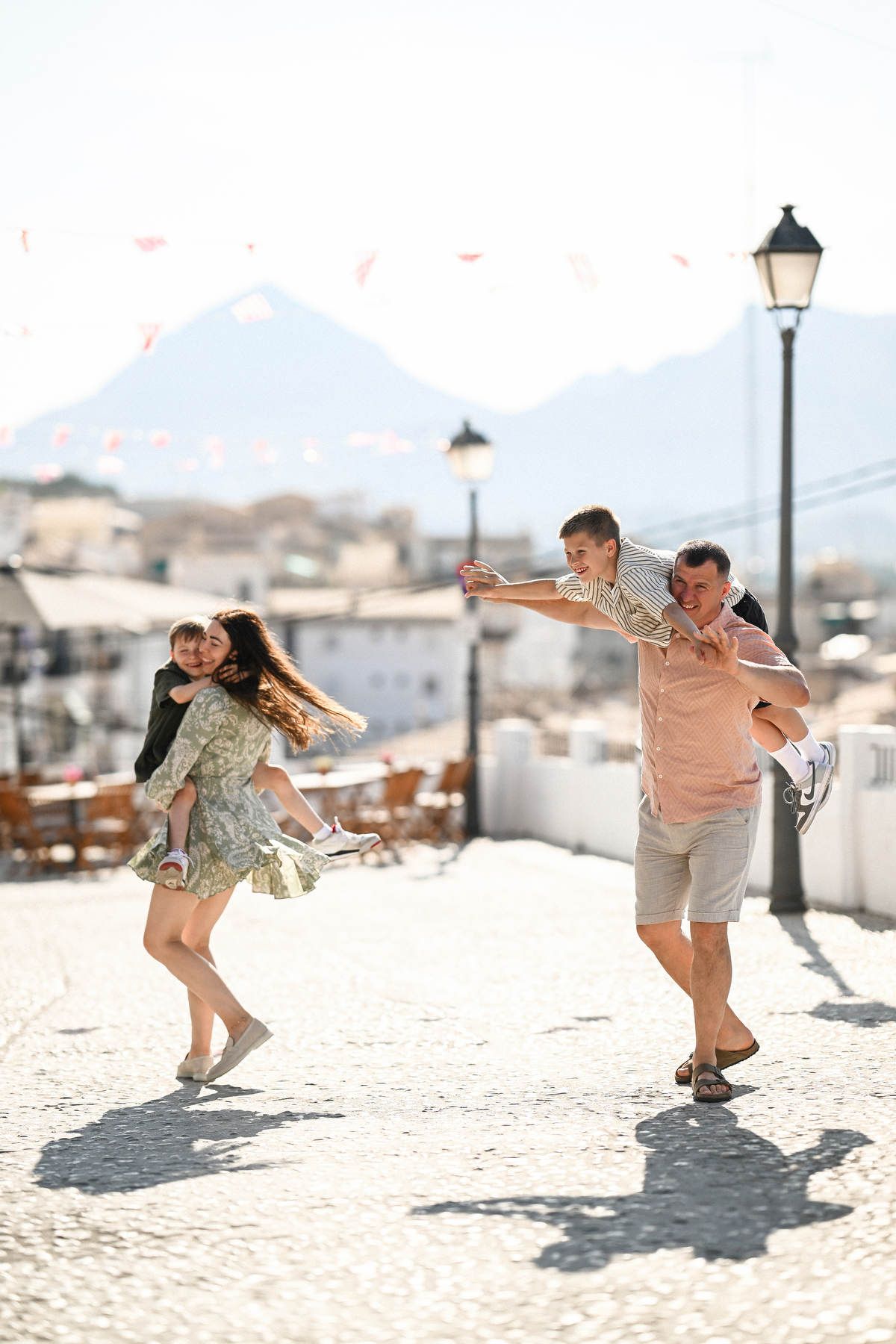 Playful moments during an Altea family session: parents carrying and spinning their kids in a sunlit historic square.