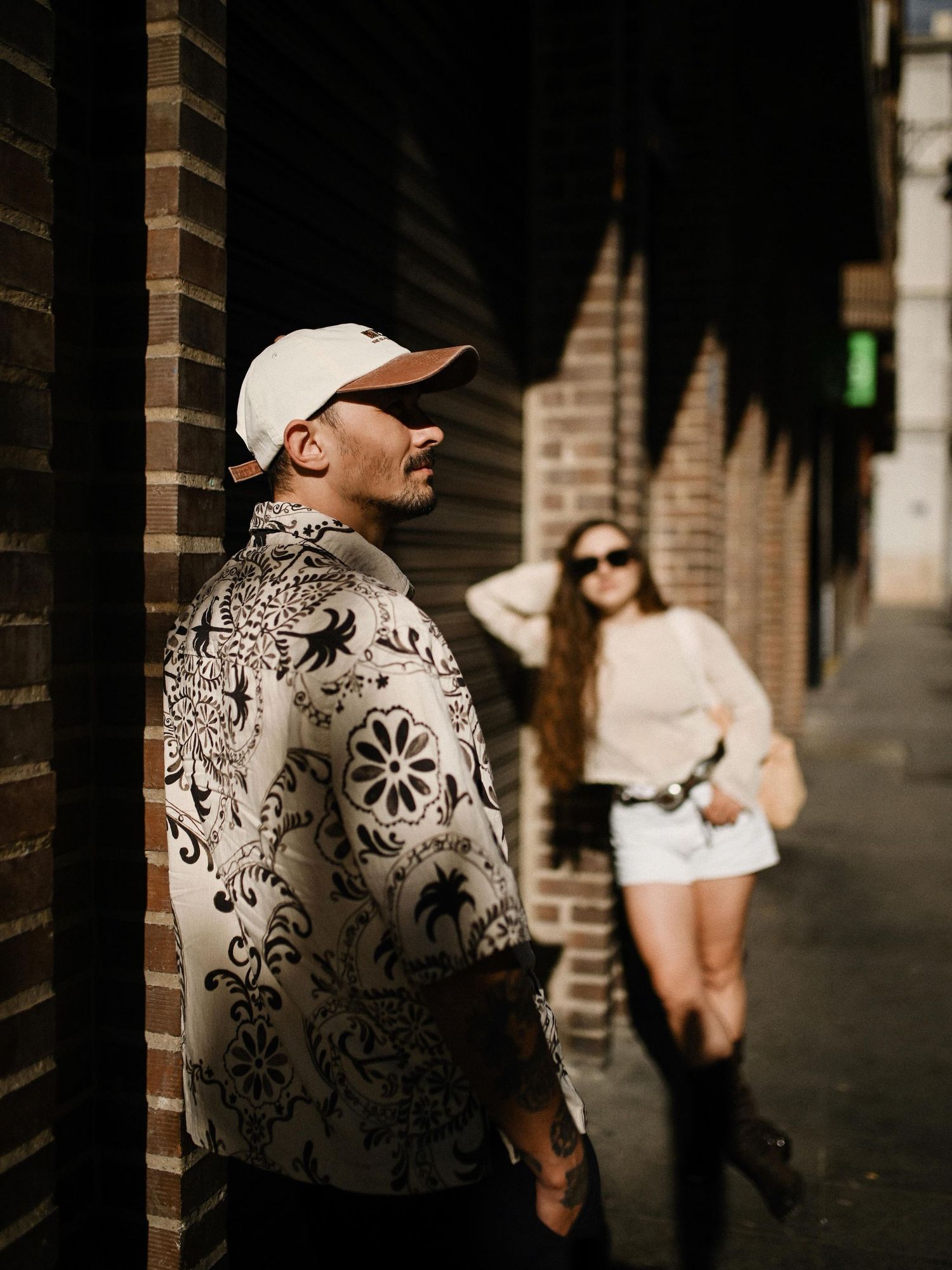 A man in a cap standing near a brick wall while his partner in sunglasses stands relaxed in the background on a city street.