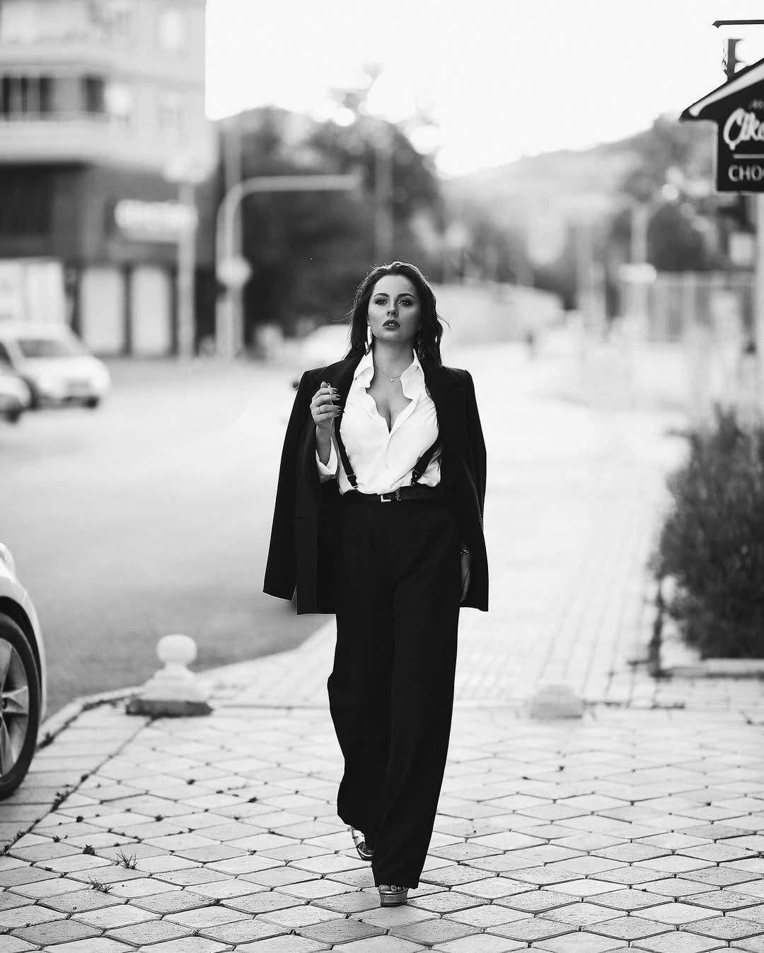A confident woman in a black blazer and white shirt walking down a city street, captured in a striking monochrome urban fashion portrait.