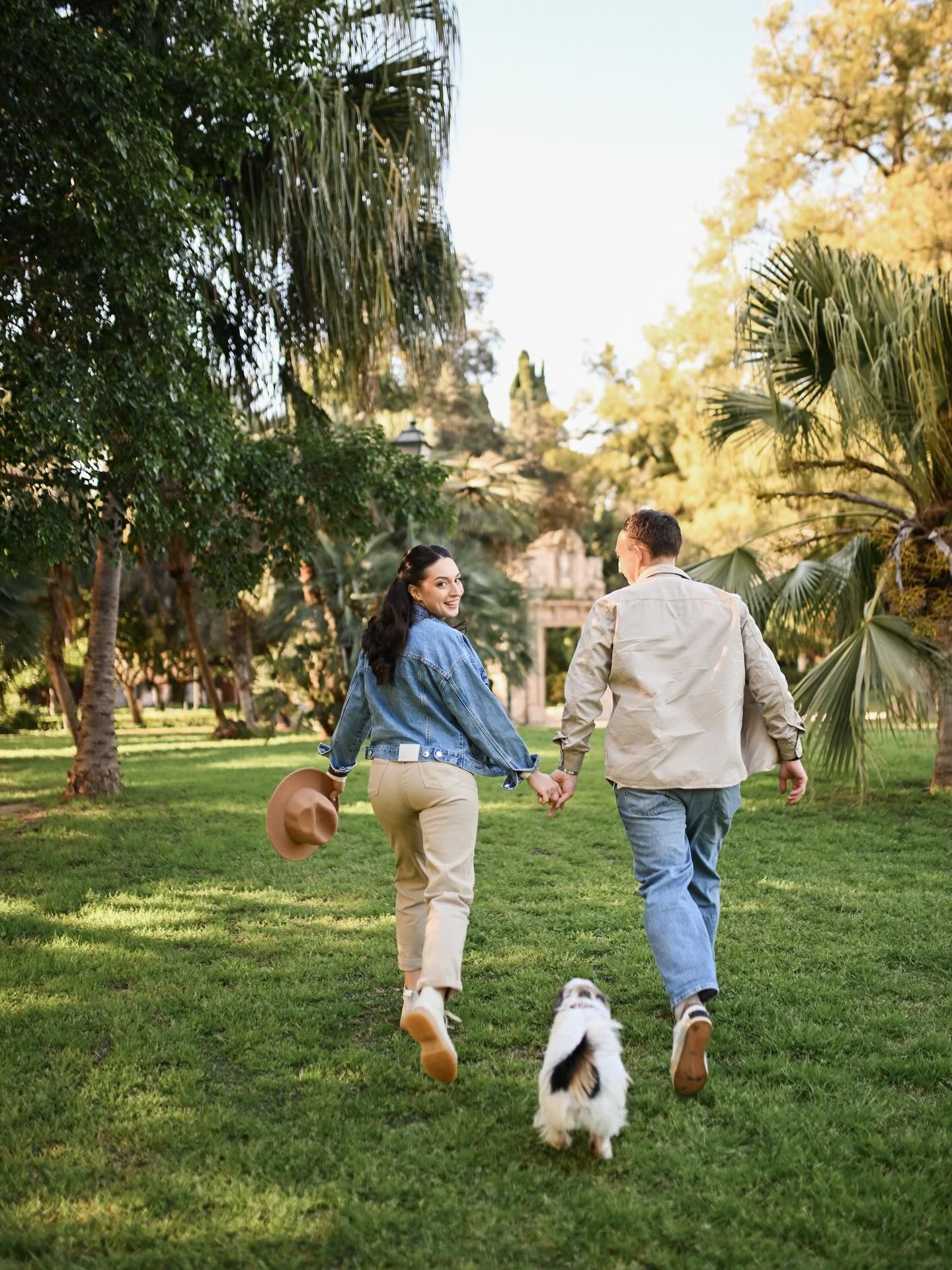 A joyful couple holding hands and running across a green lawn with their small dog, capturing a fun and active park lifestyle session.