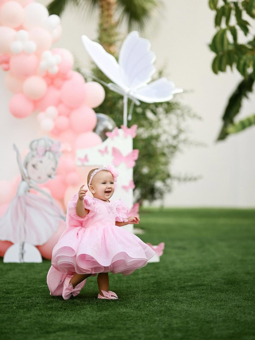 A smiling baby girl in a fluffy pink dress walking on a green lawn at an outdoor birthday celebration.