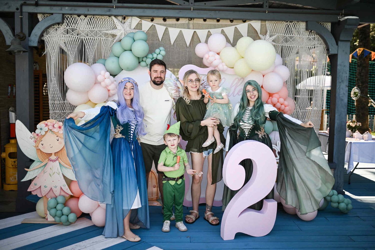A family with young children posing alongside costume fairy animators at a styled second birthday photo zone.