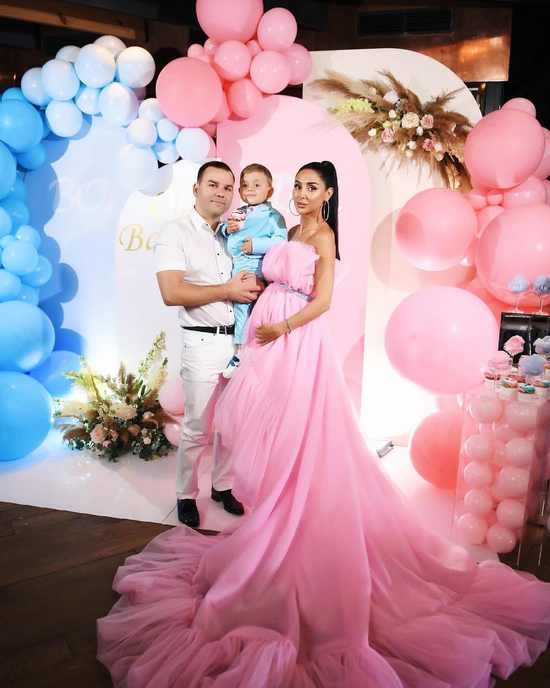 Expecting family of three posing by a pink and blue balloon arch during a beautifully styled indoor gender reveal party in Valencia.