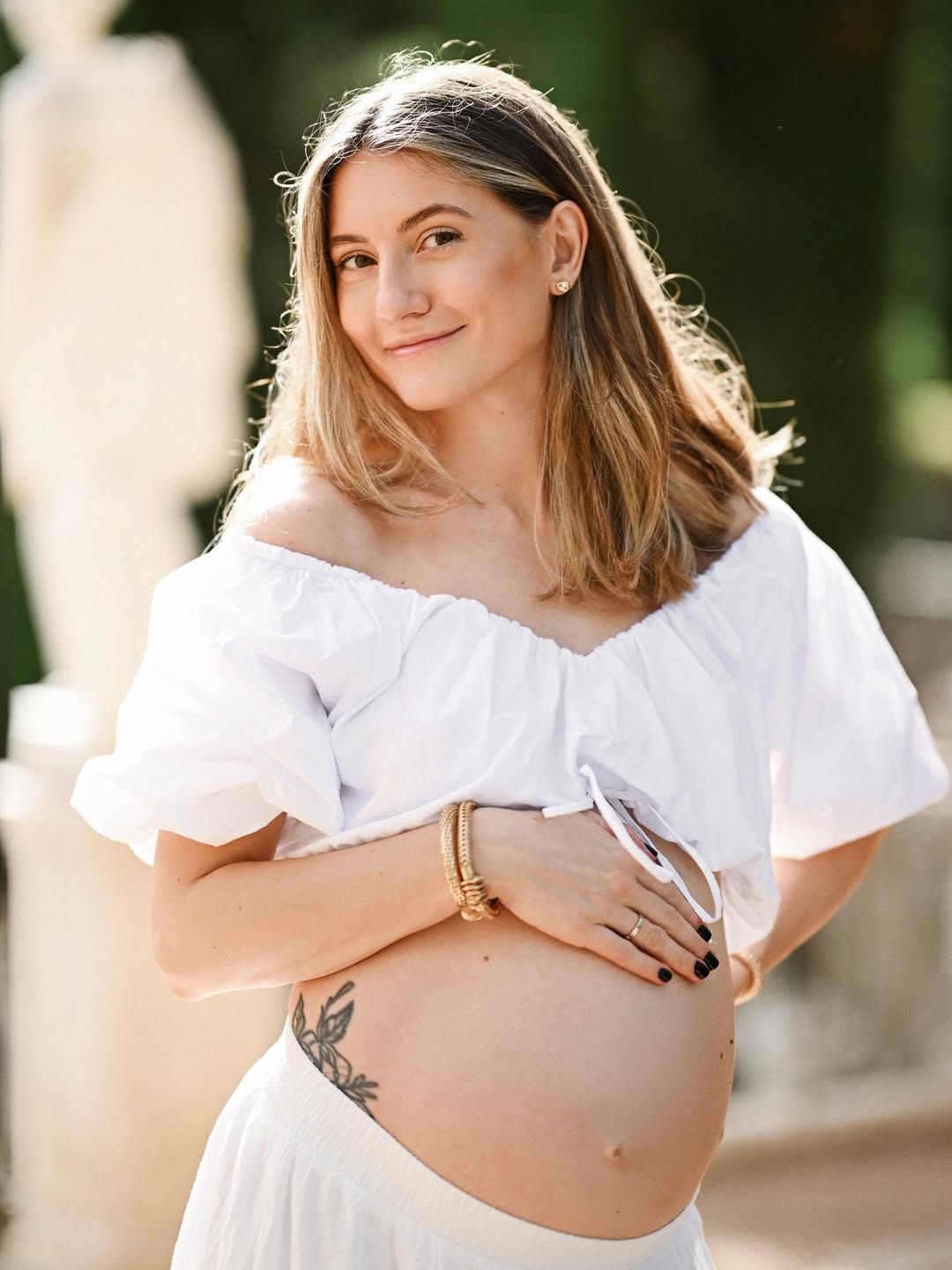Smiling pregnant woman in a white summer outfit holding her belly by a marble statue at Monforte Gardens, Valencia