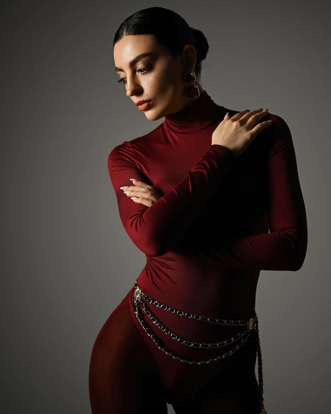 A elegant medium studio portrait of a woman with crossed arms, wearing a fitted burgundy bodysuit with a decorative silver chain belt.