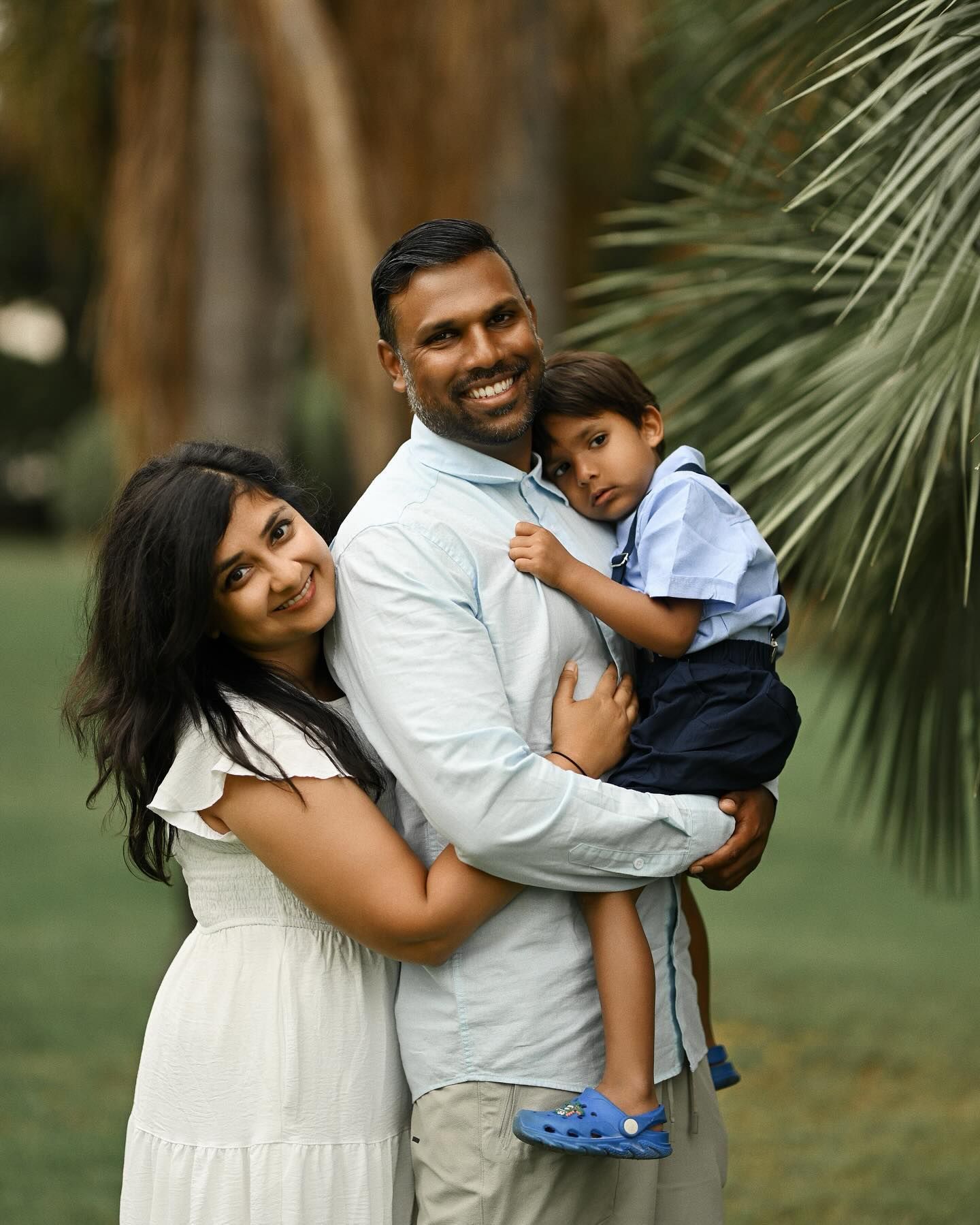 A loving family portrait in a park featuring a smiling mother and father cradling their young son, set against lush palm tree leaves.