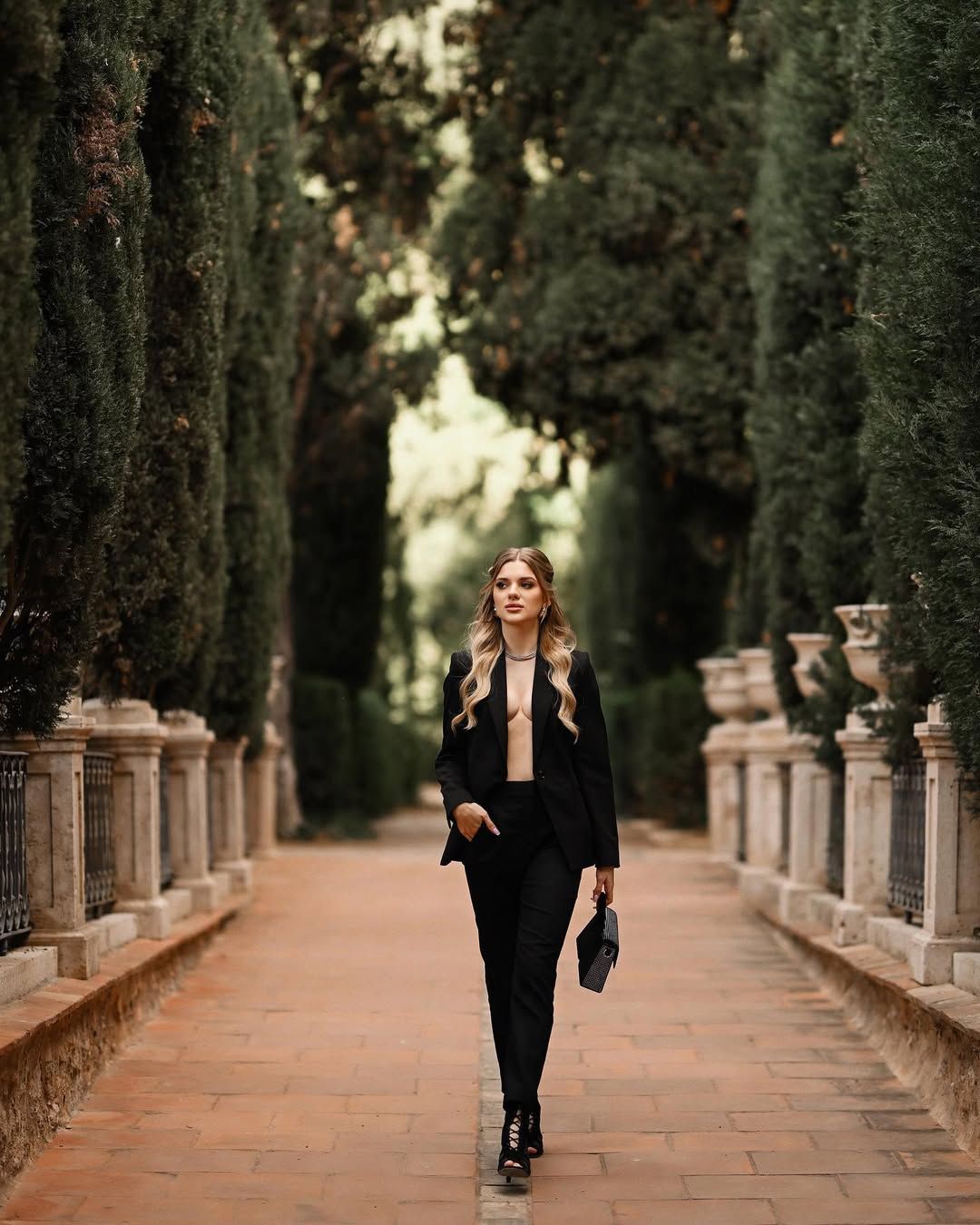 A woman in a black suit walking down a symmetrical path lined with tall green cypress trees in Valencia.