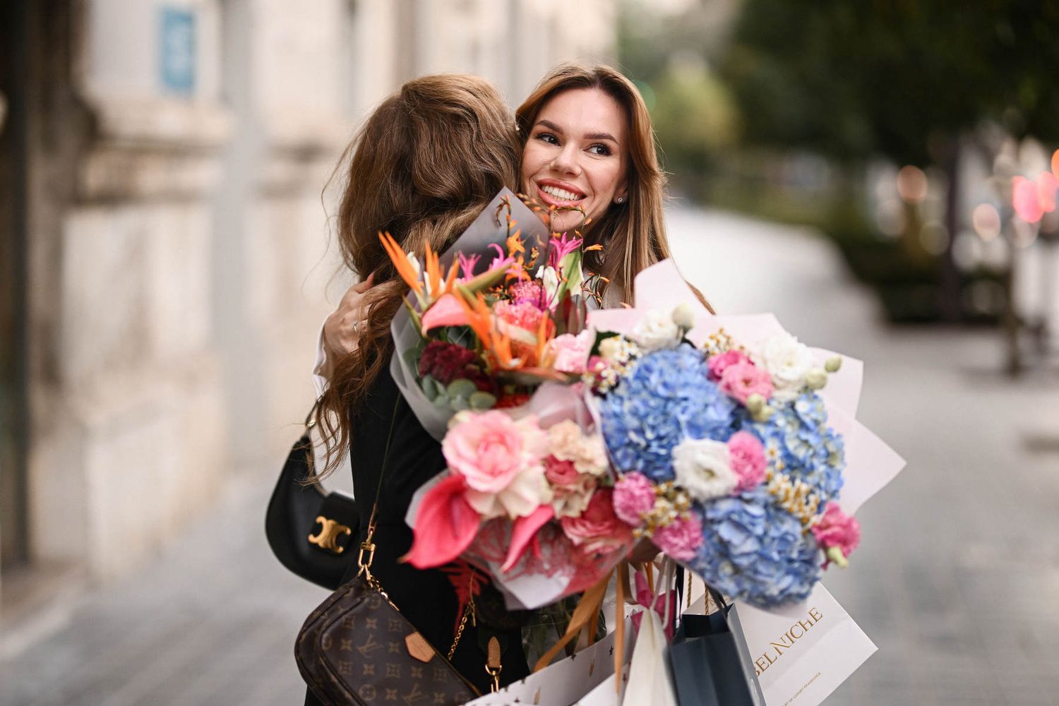 A smiling woman holding huge bouquets of pink and blue flowers while hugging a guest at a lifestyle event in Valencia.