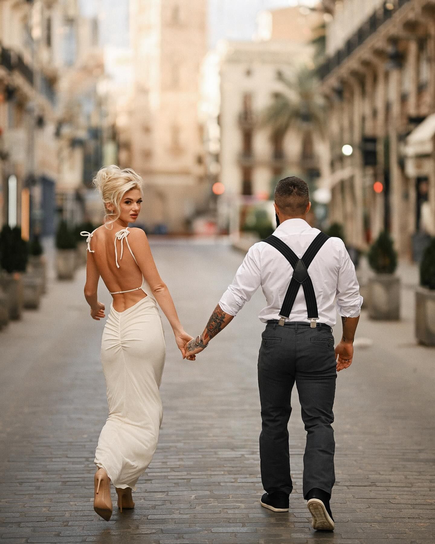 A bride in an open-back dress looking back at the camera while walking hand-in-hand with her groom down a historic Valencia street.