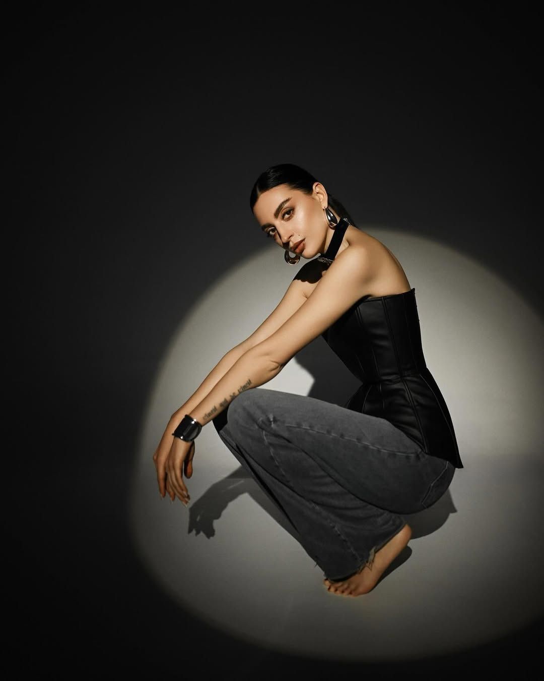 A woman in a black leather corset and grey jeans posing inside a dramatic circle of spotlight in a dark Valencia photo studio.