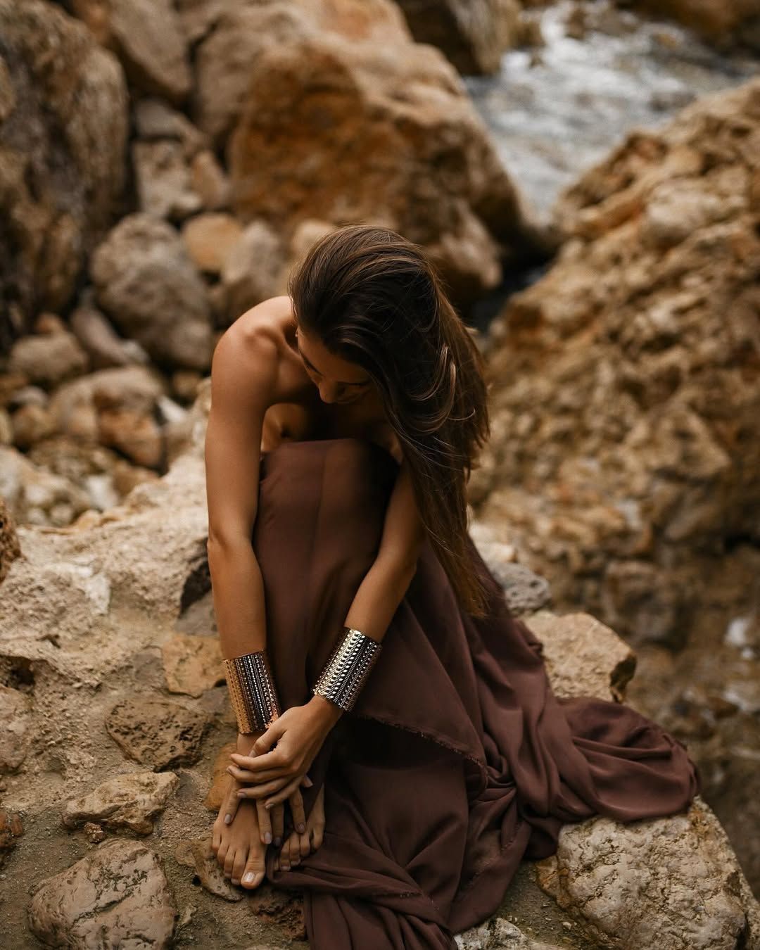 A woman with long hair wearing gold cuffs sitting on rocks during a portrait session at Cala Moraig.