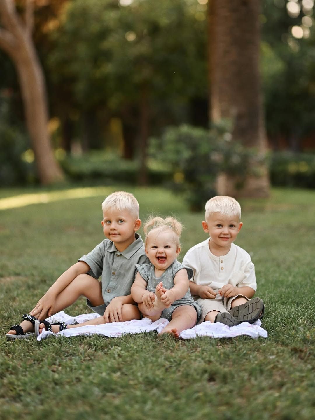 Two young boys and a baby girl sitting together on a white blanket on the grass in Turia Gardens.